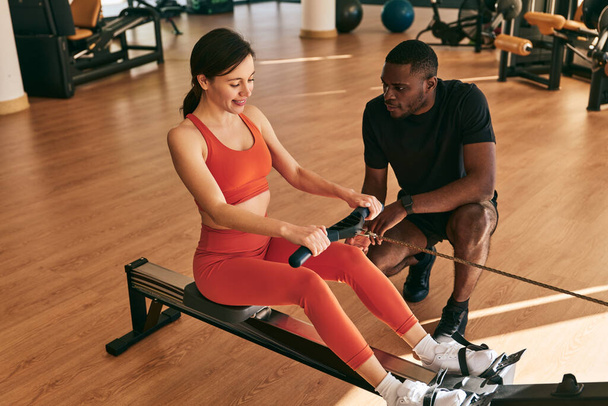 Full body high angle of young black instructor explaining technique of exercising on rowing machine, with strap to positive female in sportswear during fitness workout at gym - Foto, Bild