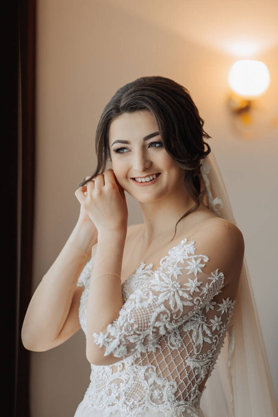 A brunette bride in a white voluminous dress with long lace sleeves, puts on her earrings, poses in her room. Great makeup. Open shoulders. Beautiful hands. Long veil. Morning of the bride. Portrait - Photo, Image