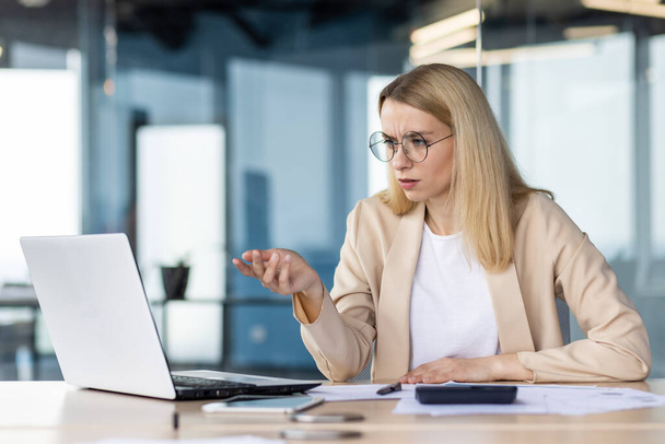 A young business woman is working in the office on a laptop, sitting at the table and talking on a video call. Upset, she holds a meeting, discusses financial problems. - 写真・画像