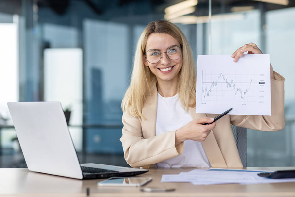 A smiling and successful young business woman is sitting at the desk in the office and showing documents with graphs to the camera. works with audits, analyses, presents diagrams of growth of work. - Foto, immagini