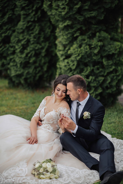Portrait of the bride in nature. A brunette bride in a white lace dress and a stylish groom in a black suit are sitting on the grass, posing, holding a bouquet. Beautiful makeup. Open shoulders. - Fotoğraf, Görsel