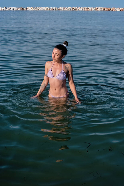Девушка с татуировками, в бикини, погруженная в морскую воду, наслаждается первым купанием летнего сезона. - Фото, изображение