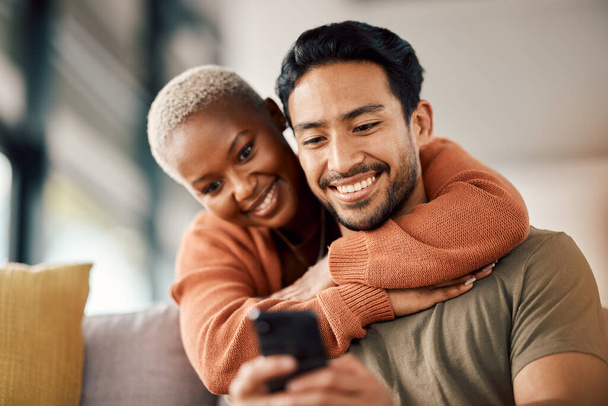 Phone, interracial couple and technology on a sofa at home reading text in a living room. Support, young people and hug together in a house sitting on a lounge couch with mobile streaming and video. - Foto, imagen