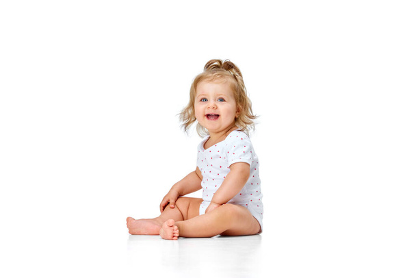 Schönes, glückliches, lächelndes Kind. Kleines Mädchen, Kleinkind, das vor weißem Studiohintergrund vor Freude und Spaß auf dem Boden sitzt. Konzept der Kindheit, Mutterschaft, Pflege, Leben, Geburt. Kopierraum für Werbung - Foto, Bild