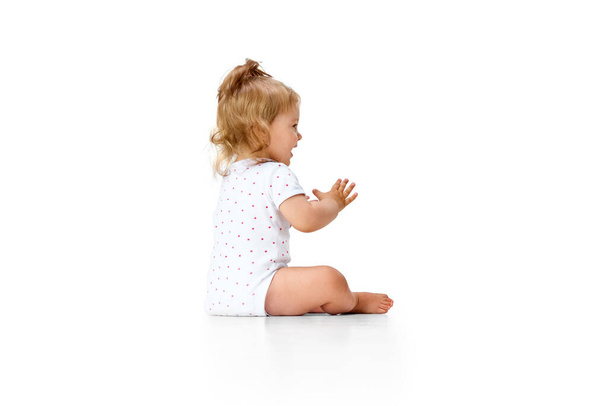 Positief, gelukkig, zorgeloos meisje kind zittend op de vloer en klappende handen in vreugde en plezier tegen witte studio achtergrond. Concept van kindertijd, moederschap, zorg, leven, geboorte. Kopieer ruimte voor advertentie - Foto, afbeelding