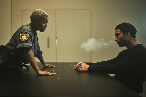 Man, smoking and detective talking in interrogation room with criminal, suspect or interview with a gangster. Police officer, questioning and statement of truth on crime and person with cigarette. - Photo, Image