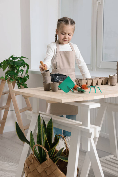 小さな女の子が部屋の木製のテーブルの泥炭鍋に土を追加します。野菜種の栽培 - 写真・画像