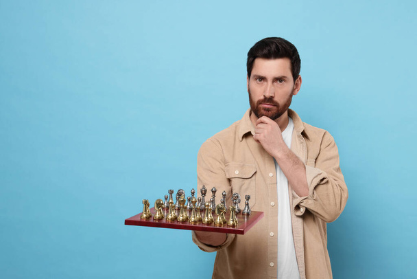 Thoughtful man holding chessboard with game pieces on light blue background. Space for text - Foto, afbeelding