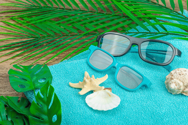 Летние каникулы. Ванное полотенце, солнцезащитные очки, морской декор. Пальмовые и чудовищные листья, деревянный фон, плоская кладка, крупным планом - Фото, изображение