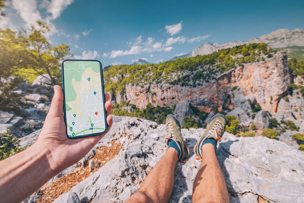 Hombre joven y deportivo en la cima de una montaña con una vista impresionante de cañón desfiladero en Turquía mira en un teléfono inteligente con una aplicación de mapa para la navegación - Foto, Imagen