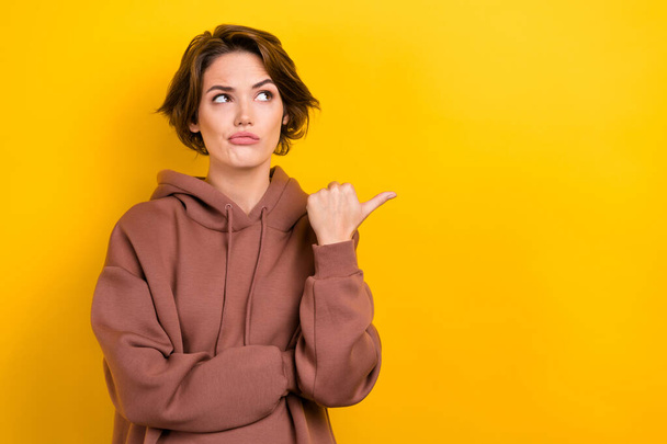 Photo of brunette hair girl indicate thumb finger empty space distrustful face wear brown hood shirt isolated yellow color background. - Foto, Imagem