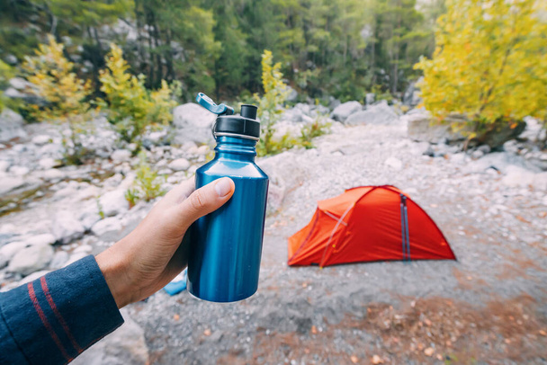 Bottle or flask with water in hand against camping tent in the woods - Photo, Image