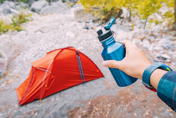Bouteille ou flacon avec de l'eau à la main contre tente de camping dans les bois - Photo, image