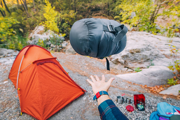 キャンプ場のテントや各種キャンプ用品などを背景に、寝袋付きの軽量・小型圧縮バッグ - 写真・画像