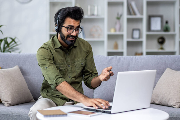Young man working from home remotely, idus using headset phone and laptop for video call and remote communication with clients, businessman sitting at home in living room on sofa. - Foto, imagen