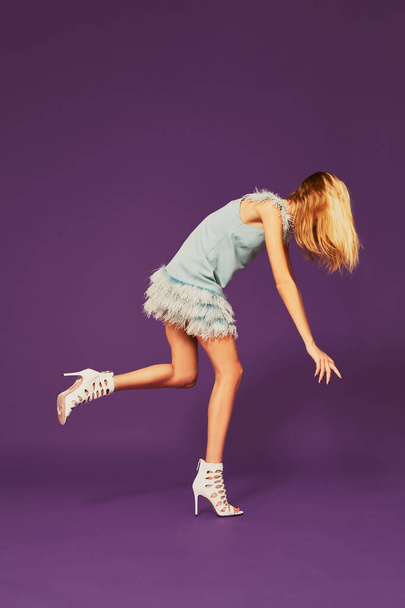 Photo of pretty young blonde girl in blue cocktail dress, trying to keep her balance. Fashion art photo shoot in studio - Foto, Imagem