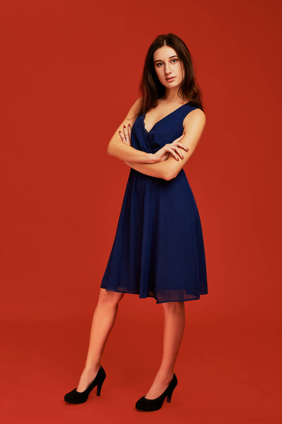 Beautiful young brunette woman in elegant blue cocktail dress and black high heels is posing for the camera. Front view. Full length studio shot on red background - Фото, изображение