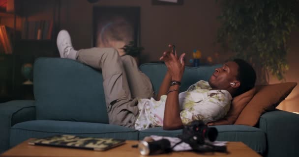 El hombre africano se reclina en el sofá, perdido en su propio mundo mientras escucha música a través de auriculares conectados a su teléfono celular. Podría ser un entusiasta de la música, disfrutando de sus canciones favoritas. - Imágenes, Vídeo