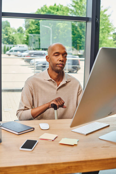 inclusion, bold african american man with myasthenia gravis, dark skinned office worker sitting with walking cane and using computer, looking at monitor, keyboard and notebook on desk  - Photo, Image
