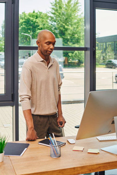синдром миастении грависа, смелый африканский бизнесмен, стоящий с тростью и смотрящий на монитор компьютера, темнокожий офисный работник в повседневной одежде, стоящий за столом, включение  - Фото, изображение