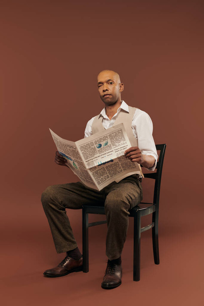 inclusion, bold african american man with myasthenia gravis syndrome, sitting on chair and reading newspaper, dark skinned black man with chronic disease on brown background - Photo, Image