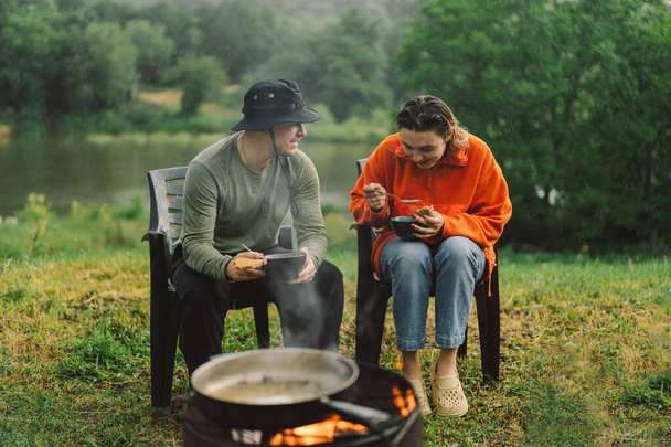 Un hombre y una mujer disfrutan de la comida junto al fuego. Campamento de senderismo con una hoguera en el bosque. Turista en recreación exterior. Estilo de vida del camping - Foto, Imagen