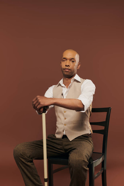 inclusion, african american man with myasthenia gravis syndrome, sitting on chair and leaning on walking cane, looking at camera, bold dark skinned man with chronic disease on brown background - Foto, afbeelding
