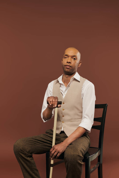 african american man with myasthenia gravis syndrome, sitting on chair and leaning on walking cane, looking at camera, bold dark skinned man with chronic disease on brown background, inclusion  - Foto, immagini
