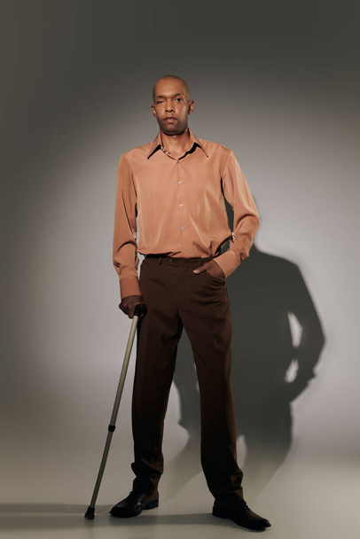 echte mensen, stoere Afro-Amerikaanse man met myasthenia gravis staande met wandelstok op grijze achtergrond, hand in zak, pose, donkere huid persoon in shirt, diversiteit en inclusie, volledige lengte  - Foto, afbeelding