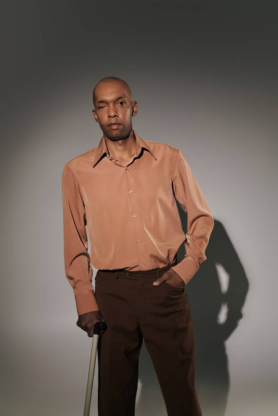 реальні люди, сміливий афроамериканський чоловік з міастенією, що стоїть з ходячою тростиною на сірому фоні, рука в кишені, поза, темношкіра людина в сорочці, різноманітність та включення  - Фото, зображення