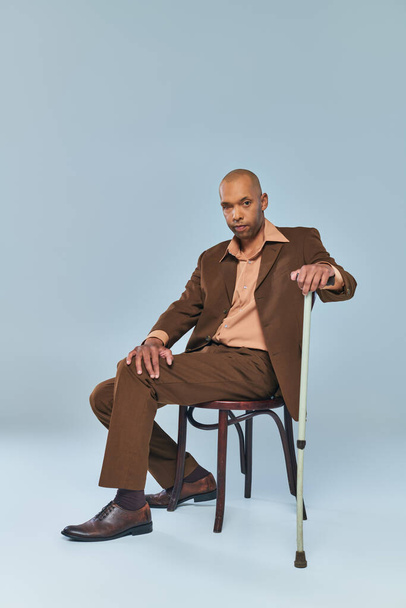 etniczny, pełnoetniczny, odważny, afrykański mężczyzna z miastenią Gravis siedzący na krześle na szarym tle, ciemnoskóry człowiek w garniturze opierający się na chodzącej lasce, różnorodności i integracji  - Zdjęcie, obraz