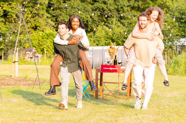An engaging image capturing a moment of joy and competition as two multicultural couples participate in a lively piggyback race during a garden party. Their expressions and camaraderie exude fun - Photo, Image
