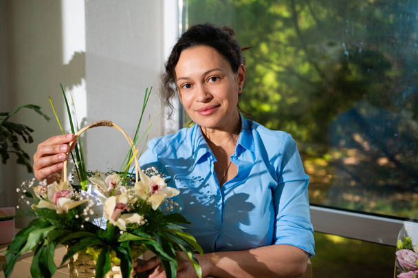 Zelfverzekerde multi-etnische jonge vrouw, startupper, ondernemer, glimlachend kijkend naar de camera, werkend in een bloemenwinkel, zittend aan een tafel met kamerplanten en bloemen. Mensen. Lifestyle. Hobby - Foto, afbeelding