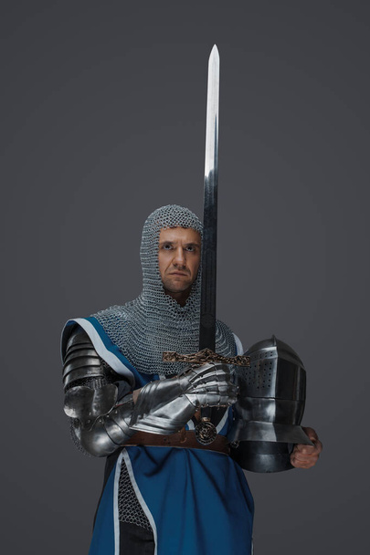 Orgoglioso cavaliere regge la spada e l'elmo, indossa un soprabito blu, un cappotto a catena e un'armatura medievale, sullo sfondo grigio - Foto, immagini