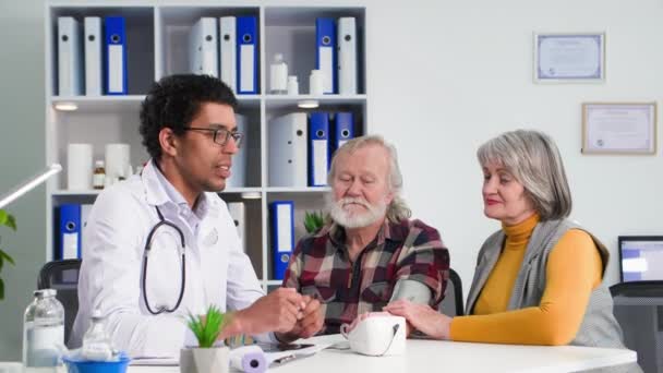 ricevimento di pazienti, una coppia anziana consulta sullo stato di salute con il giovane medico nero professionale nello studio medico - Filmati, video