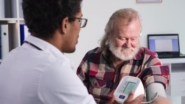 медицинское обслуживание, пожилой человек на приеме у молодого черного врача измеряет давление тонометром в медицинском кабинете - Кадры, видео