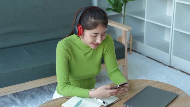 Χαλαρή Ασιάτισσα που φοράει ακουστικά χρησιμοποιώντας smart phone surfing στα μέσα κοινωνικής δικτύωσης, τσεκάροντας ειδήσεις, ακούγοντας μουσική ή στέλνοντας μηνύματα που κάθονται στον καναπέ. Η Χιλιετής κυρία περνάει χρόνο στο σπίτι. Υψηλή ποιότητα - Πλάνα, βίντεο