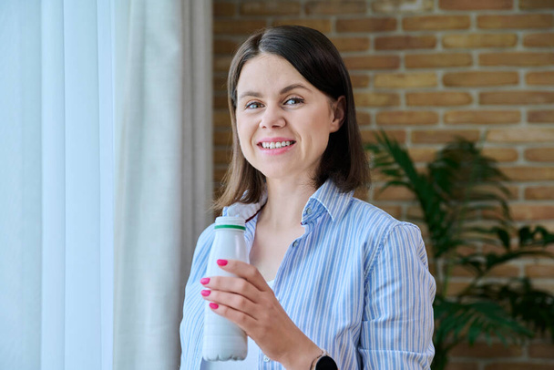 Young woman drinking fermented milk product from bottle, at home near window, healthy breakfast. Dairy vitamin products, yogurt, kefir, diet, healthy eating concept - Zdjęcie, obraz