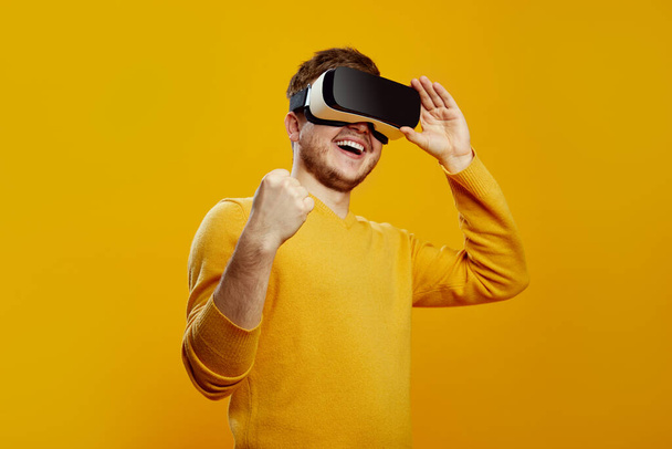 Smiling excited young man wearing yellow sweater watching in vr headset pc gadget doing winner gesture, celebrating against orange background - Фото, изображение