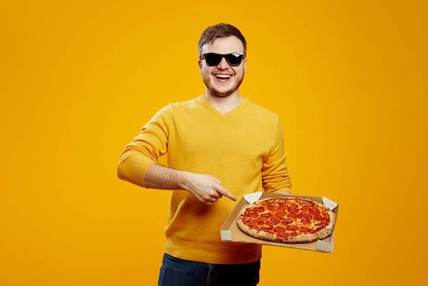 Hombre barbudo joven guapo en suéter amarillo y gafas de sol sosteniendo y señalando la pizza en caja plana de cartón aislado sobre fondo naranja - Foto, imagen