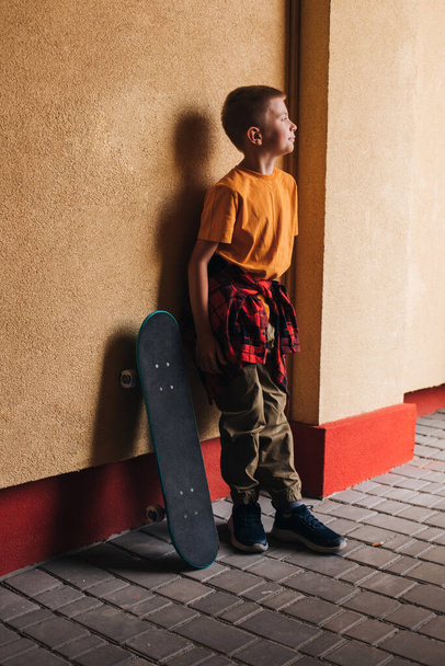 Atividades ao ar livre para crianças. Um menino alegre em roupas brilhantes com um skate contra o pano de fundo de uma parede colorida. Um estudante caucasiano a passear. Vista frontal - Foto, Imagem