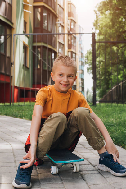 На скейтборде сидит веселый мальчик в яркой одежде. Белый школьник на прогулке. Вид спереди - Фото, изображение