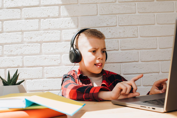 Хлопчик шкільного віку в чорних навушниках сидить за столом з ноутбуком. Діти і гаджети. Вид спереду
 - Фото, зображення