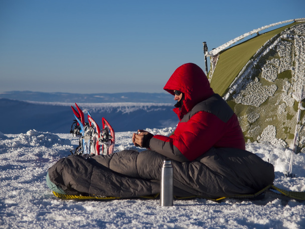 A man sits in a sleeping bag near the tent and snowshoes. - Photo, Image