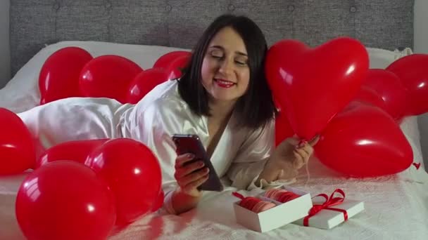 Woman lying and using her phone between red balloons on bed. Smiling woman with a lot of air balloons and gift box with macarons. Happy birthday anniversary. Red decoration for wedding, Valentines Day - Filmati, video
