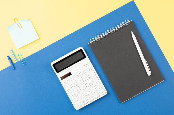 Spiral open notebook with pen and calculator on two tone background. School table concept. Office workplace. Top view, flat lay. - Photo, Image