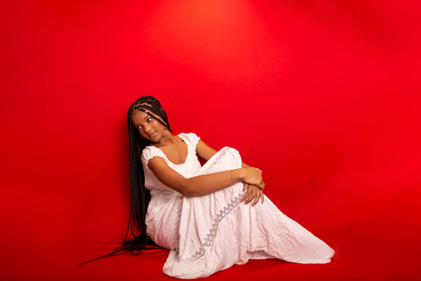 Bella giovane donna nera seduta con calma indossando abiti bianchi con lunghe trecce. Isolato su sfondo rosso - Foto, immagini