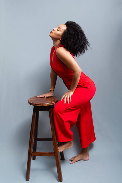 Young woman, standing, wearing a red outfit with curly hair posing next to a wooden stool. Isolated on light gray background. - Foto, Imagen