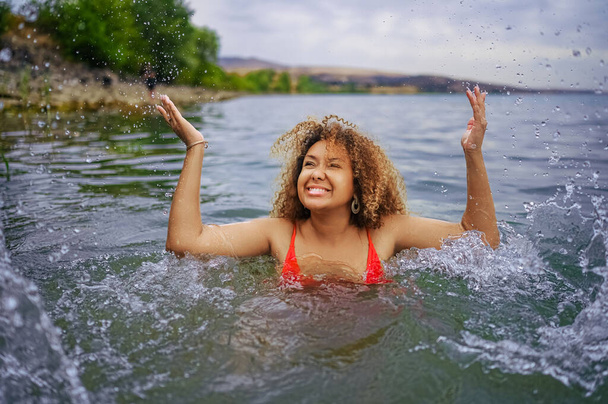 Atraente feliz plus size Africano americano preto mulher gosta de nadar se divertindo com salpicos de água na água do lago do mar em tempo nublado ao ar livre - Foto, Imagem