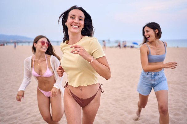 Group of three beautiful young and attractive women running happy along beach having fun. Caucasian girls enjoying summer vacation outdoors. Spanish friends people having fun weekend holiday. - 写真・画像